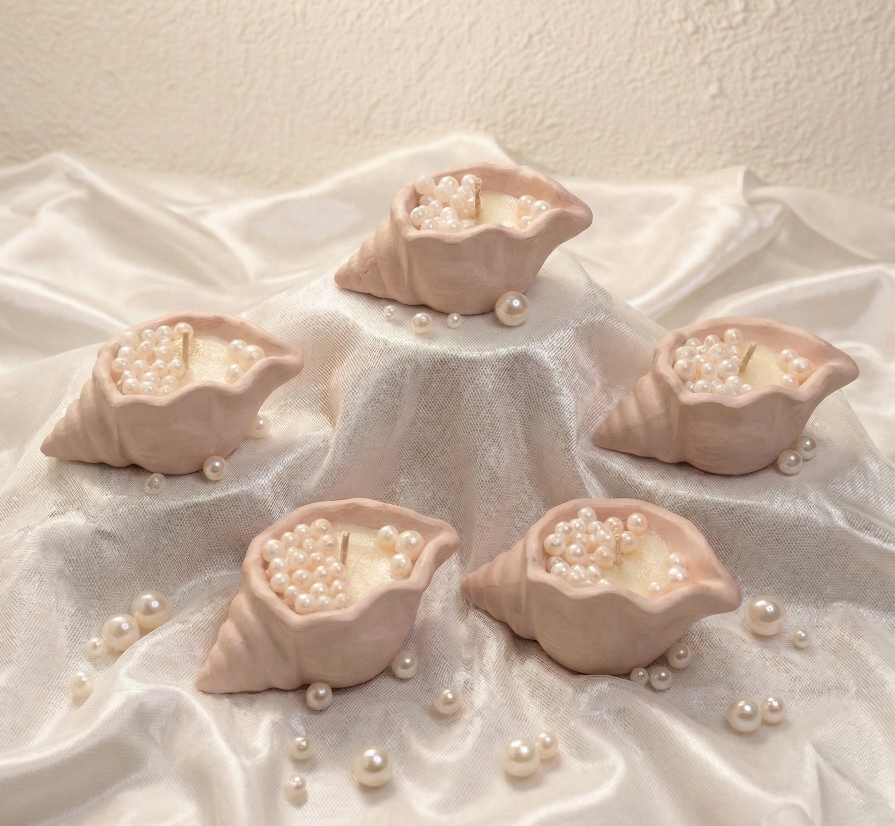 Five handcrafted blush shell candles filled with white wax pearls arranged on a white silk fabric with scattered pearls.